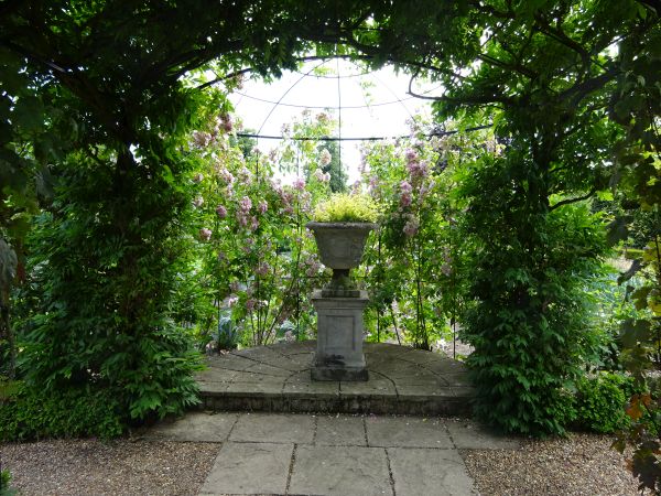 Stone urn with plants on stone plinth see through covered walkway.