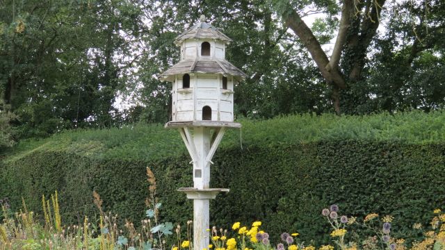 White dove house on pole with grass bank and trees behind.